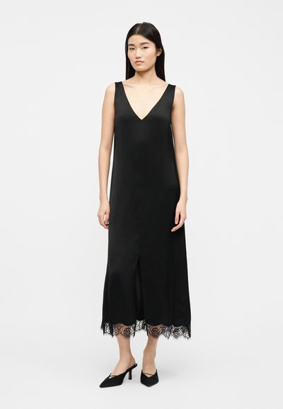 Γυναίκα που στέκεται φορώντας αμάνικο μαύρο μίντι φόρεμα με V-λαιμόκοψη και τελείωμα με δαντέλα, συνδυασμένο με μαύρα μυτερά mule με τακούνι.