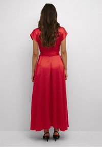 Kvinna med långt brunt hår som bär en golvlång röd klänning med ett subtilt mönster, stående mot en enkel ljusgrå bakgrund.