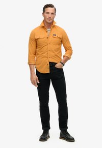Orange skjorte med knapper og to brystlommer, båret over en grå skjorte. Sorte jeans og snørebåndstøvler fuldender looket.