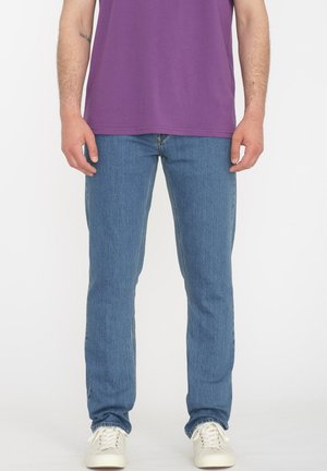 Light blue denim jeans with a slim fit, featuring five pockets and a subtle vertical texture, paired with a purple t-shirt and white sneakers.