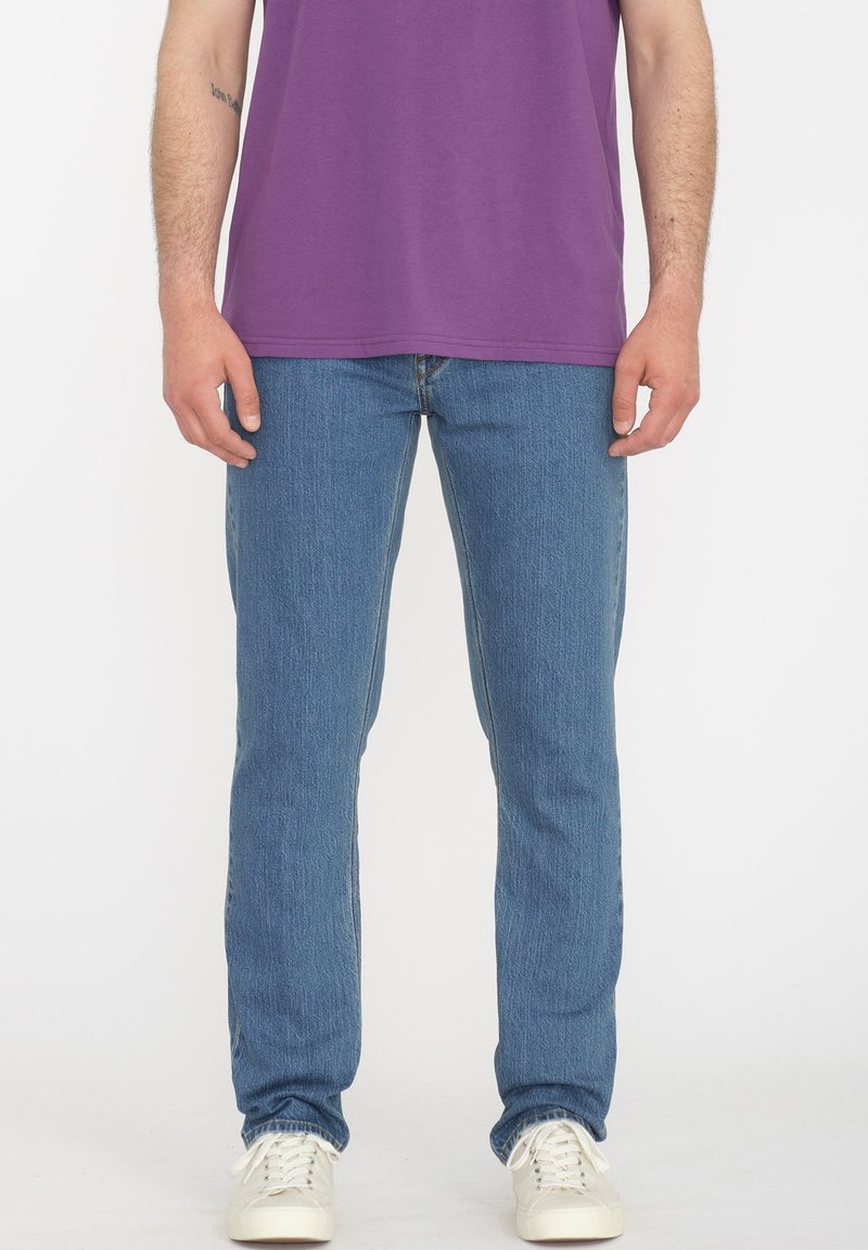 Jeans de denim azul claro con un corte ajustado, que cuentan con cinco bolsillos y una textura vertical sutil, combinados con una camiseta morada y zapatillas blancas.
