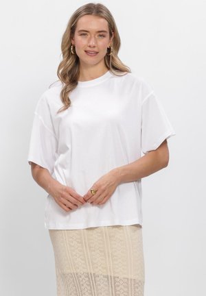Vrouw met golvend blond haar die gouden creolen draagt, een eenvoudig wit oversized T-shirt en een beige rok met patroon tegen een witte achtergrond.
