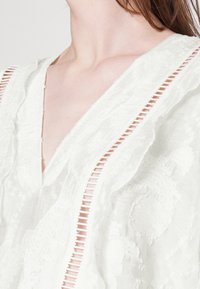 Blusa bianca con scollatura a V profonda, caratterizzata da un tessuto testurizzato e strisce ritagliate verticali lungo i bordi. Design leggero e arioso.