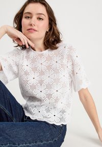 Weißes Spitzenoberteil mit Blumenmustern und kurzen Ärmeln, mit gewellten Kanten. Über dunkelblauen Jeans getragen, die das strukturierte Design hervorheben.