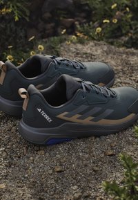 Dark green athletic shoes feature a smooth textile upper, brown accents, cushioned soles, and multicolored laces, designed for outdoor use.
