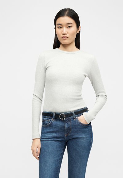 Femme aux cheveux noirs raides portant un pull à manches longues gris clair côtelé, un jean bleu et une ceinture noire avec une boucle anneau argentée.