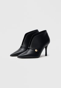 Μαύρα δερμάτινα μποτάκια μέχρι τον αστράγαλο με μυτερές μύτες, λεπτούς τακούνια και χρυσό κουμπί σε λουράκι πάνω από το παπούτσι. Λείο υφή.