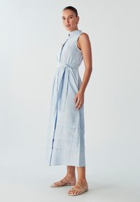 Šviesiai mėlyna, be rankovių midi suknelė pagaminta iš audinio su mygtukų užsegimu, liemeniu, keleto sluoksnių dizainu ir šonine skeltuku apačioje. Suderinta su plokščiomis šlepetėmis.