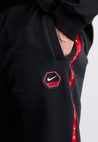 Sorte joggebukser med et hexagonalt Nike-merke og en rød aksentstripe. Stoffet har en glatt tekstur og en avslappet passform med sidelommer.
