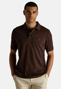 Brūna polo krekls, kas izgatavots no mīksta kokvilnas, ar apkakli, īsām piedurknēm un mazu sarkanu logotipu uz krūtīm. Kombinēts ar gaišas krāsas biksēm.