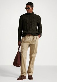 Χακί chinos, σκούρος πράσινος πουλόβερ με πλεκτό σχέδιο και ψηλό γιακά, καφέ δερμάτινη τσάντα και καφέ παπούτσια. Η εμφάνιση είναι casual με καθορισμένες υφές και σχήματα.