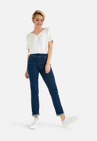 Blusa bianca a maniche corte, vestibilità rilassata, abbinata a jeans scuri a maniche risvolte. Il modello indossa sneakers bianche, mostrando un outfit casual.