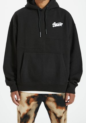 Personne portant un sweat à capuche noir avec un logo blanc sur la poitrine et un pantalon tie-dye noir, marron et beige.