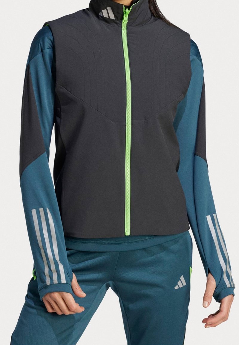 Femme portant un gilet noir sans manches avec une fermeture éclair vert néon sur un haut bleu à manches longues et un pantalon bleu assorti avec des bandes blanches sur les manches.