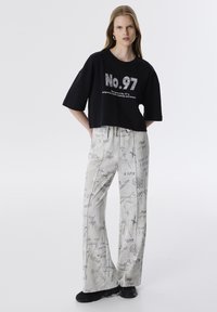 Zwarte cropped t-shirt met het grafische ontwerp "No. 97". Lichtgrijze wijde broek met verschillende handgeschreven patronen. Zwarte schoenen.