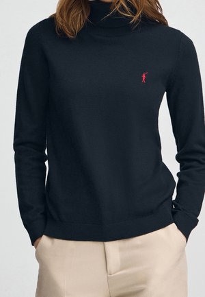Femme portant un pull à col roulé noir avec un petit logo de joueur de polo rouge sur la poitrine et un pantalon beige, les mains dans les poches.