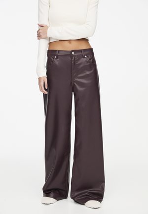 Person wearing high-waist wide-leg dark brown leather pants with button and pocket details, paired with a cropped long-sleeve white top.