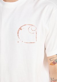 White cotton t-shirt with a front pocket and a brown circular logo design. Short sleeves, crew neckline, and soft texture.