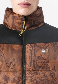 Chaqueta acolchada con un patrón floral marrón, con una sección superior negra, cierre de cremallera y un cordón amarillo en el cuello.