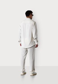 Weißes Leinenhemd mit langen Ärmeln, lockerer Passform und abgerundetem Saum; kombiniert mit passenden Leinenhosen und lässigen weißen Sneakers.