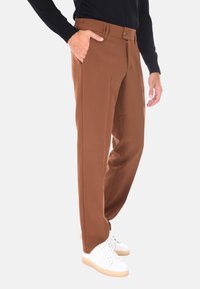 Uomo che indossa pantaloni marroni su misura, una maglietta nera a maniche lunghe e sneakers bianche, in piedi con una mano in tasca su uno sfondo chiaro.