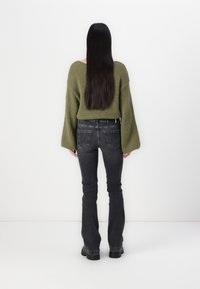 Femme avec de longs cheveux noirs raides, portant un pull vert olive et un jean noir, se tenant face à un mur blanc.
