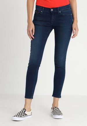 Jeans skinny de mezclilla azul oscuro con talle medio, que presentan bolsillos estándar y detalles de costura sutiles, combinados con zapatos slip-on a cuadros en blanco y negro.