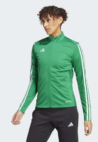 Groene sportieve jas met witte strepen op de mouwen, hoge kraag, volledige rits en ademend materiaal, gecombineerd met zwarte broek.