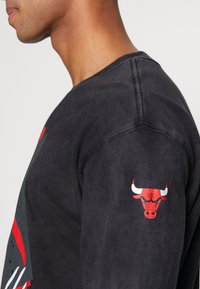 Svart långärmad sweatshirt med en grafisk bild av en röd och vit tjur på ärmen och en stiliserad grafik över framsidan. Mjuk textur.