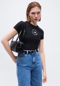 Νεαρή γυναίκα που φορά σφιχτό μαύρο μπλουζάκι με κυκλικό λογότυπο, τζιν παντελόνι ψηλόμεσο μπλε, μαύρη ζώνη, σκουλαρίκια κρίκους και κρατά μαύρη δερμάτινη τσάντα ώμου.