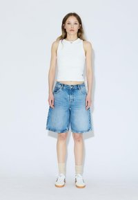 Haut sans manches blanc, shorts en denim bleu, et baskets blanches. Les shorts sont de longueur genoux avec une coupe décontractée et un design classique à cinq poches.