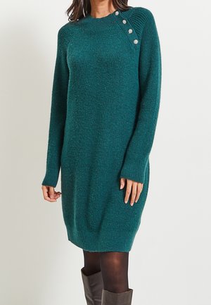 Robe-pull en maille bleu sarcelle à manches longues, avec des boutons décoratifs argentés sur l'épaule, portée avec des collants noirs et des bottes marron montant au genou.