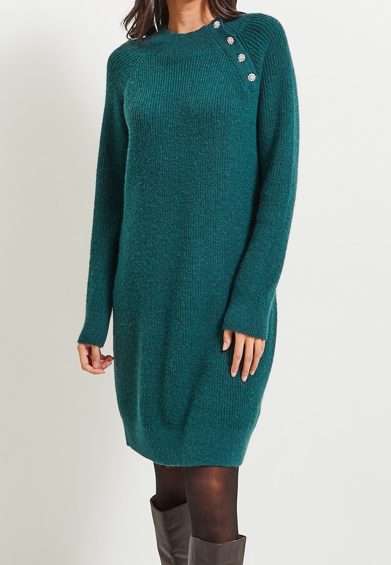 Robe-pull en maille bleu sarcelle à manches longues, avec des boutons décoratifs argentés sur l'épaule, portée avec des collants noirs et des bottes marron montant au genou.