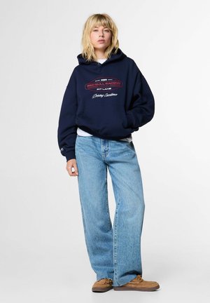 Blonde vrouw met een navy blauwe Red Bull Racing hoodie, lichtblauwe wijde jeans en bruine mocassins, staand tegen een effen witte achtergrond.