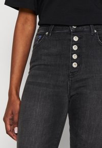 Vaqueros negros de cintura alta con un cierre frontal de botones, cinco botones plateados y una textura clásica de denim. Presenta bolsillos laterales y presillas para el cinturón.