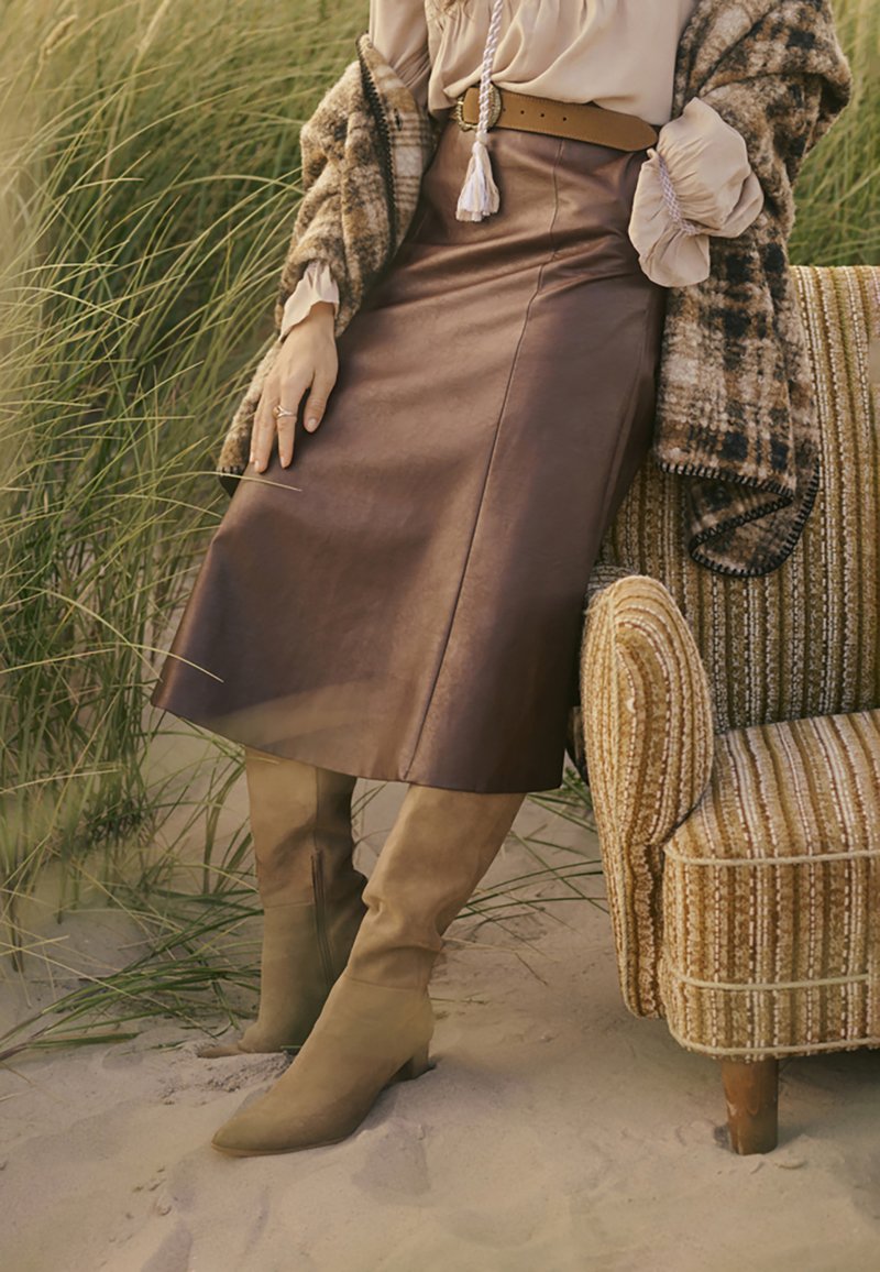 Jupe en cuir marron au design ajusté, assortie à des bottes beiges hautes. La tenue est complétée par un châle en tricot texturé beige et marron.