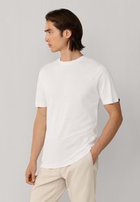 Jeune homme tourné vers la gauche portant un t-shirt blanc ajusté à manches courtes et un pantalon beige, une main dans la poche, sur un fond uni.
