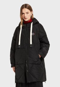 Sort dunjakke med diamantformet quiltet mønster, lynlås foran, hvide snore, hætte og logo-patch. Tekstureret stof og overdimensioneret pasform.