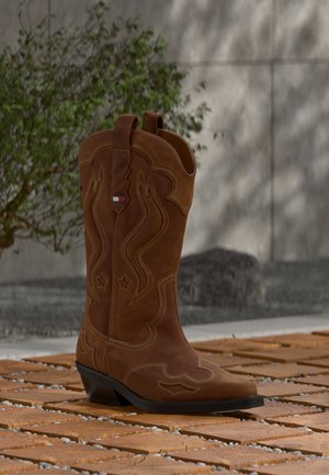 Stivale da cowboy in camoscio marrone con cuciture decorative e un emblema con bandiera sul gambale, posato su un pavimento in doghe di legno all'aperto.