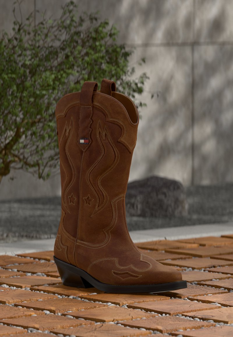 Botte de cowboy en daim marron avec des coutures décoratives et un emblème de drapeau sur la tige, posée sur un sol en lattes de bois à l'extérieur.