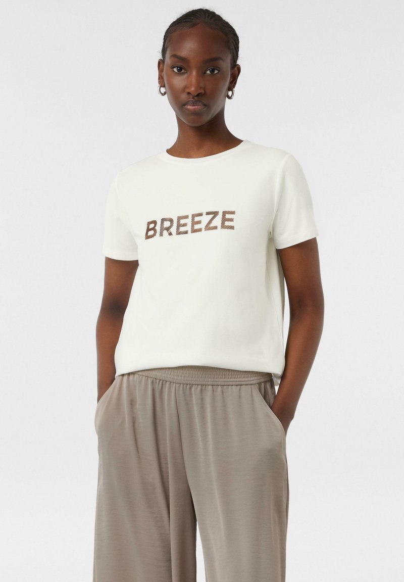 Helles beige T-Shirt mit kurzen Ärmeln und braunem Text "BREEZE" auf der Vorderseite, kombiniert mit locker sitzenden taupefarbenen Hosen mit Seitentaschen.