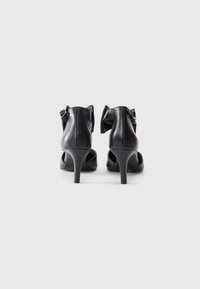 Black leather high-heeled shoes featuring a curved back, ankle bow detail, and a slim stiletto heel with a smooth texture.