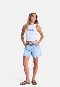 Witte crop top met turquoise bloemenprint, lichtblauwe denimshorts en bruine slidesandalen. Model poseert met handen in de zakken.