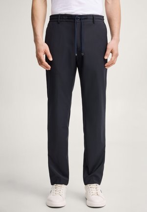 Homme portant un pantalon bleu marine à cordon, un t-shirt blanc et des baskets blanches, debout devant un fond uni clair.