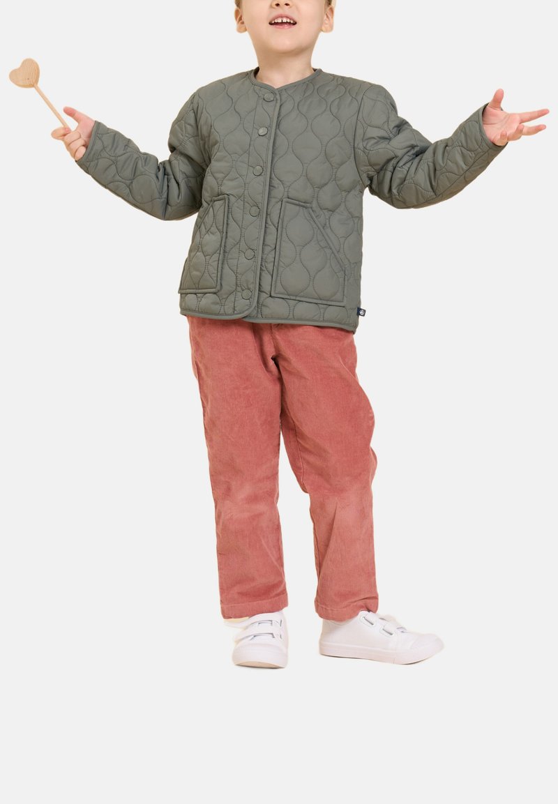 Veste matelassée verte avec un col rond et des boutons-pression, associée à un pantalon en velours côtelé rose et des baskets blanches. Tige en bois en forme de cœur tenue.