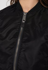 Czarna kurtka bomberka wykonana z gładkiego materiału, z przednim zamkiem błyskawicznym, prążkowanym kołnierzem i wyhaftowanym logo po lewej stronie.