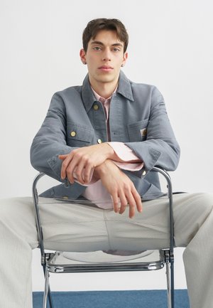 Joven con pendientes sentado de espaldas en una silla, lleva chaqueta gris, camisa rosa y pantalones de color claro, sobre un fondo liso.