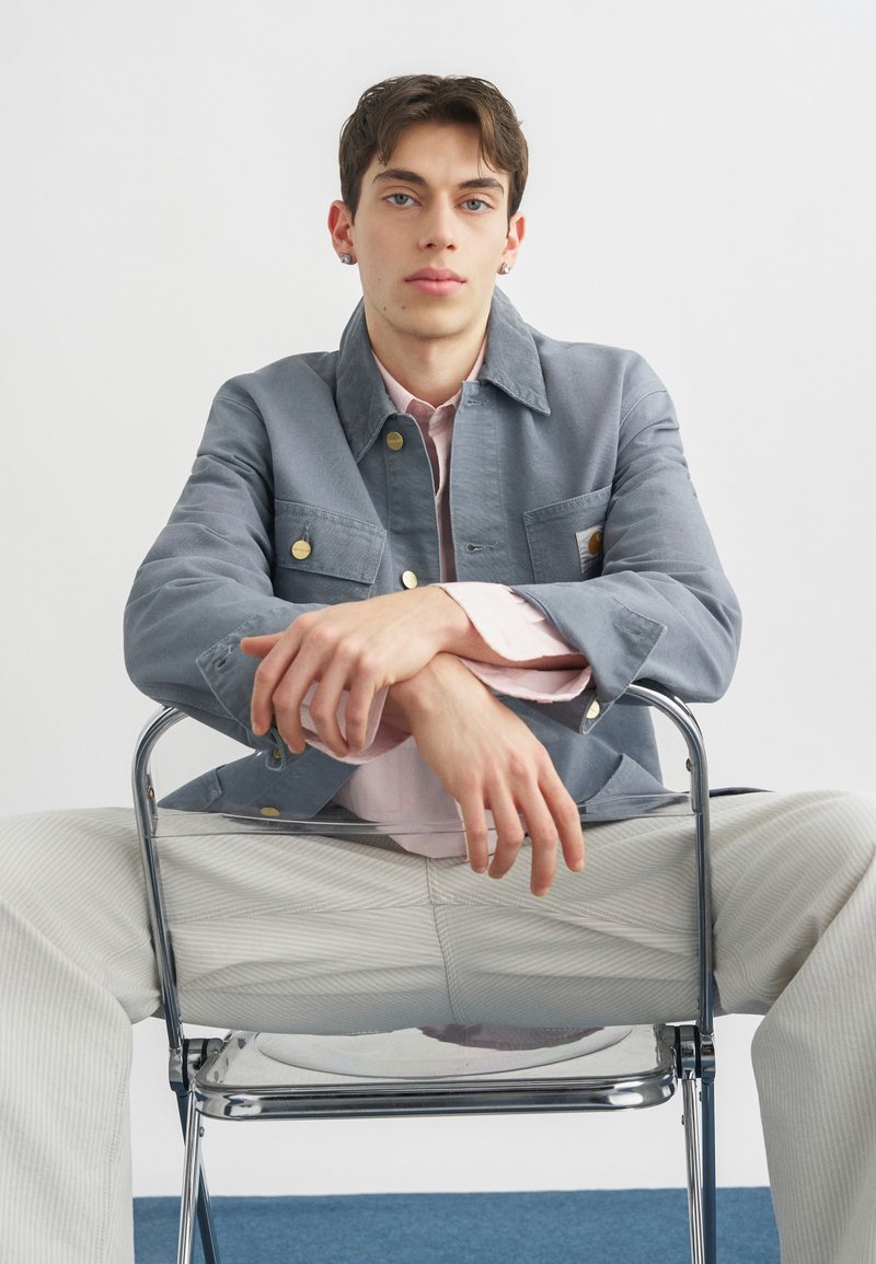 Giovane uomo con orecchini seduto su una sedia al contrario, indossa giacca grigia, camicia rosa e pantaloni chiari su sfondo semplice.