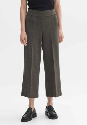 Femme portant un pantalon marron en chevrons, taille haute, jambes larges et coupé au-dessus de la cheville, et des mocassins noirs à enfiler, debout sur un fond blanc.
