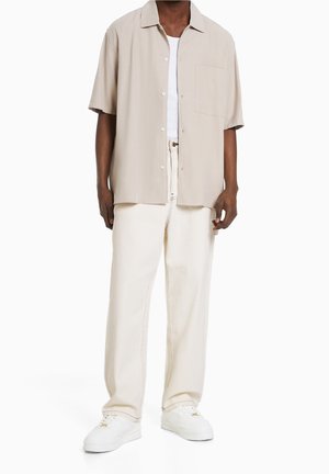 Chemise beige à manches courtes avec col pointu, fermeture par boutons, poche poitrine, assortie à un pantalon blanc et des baskets blanches, mettant en avant un style décontracté.